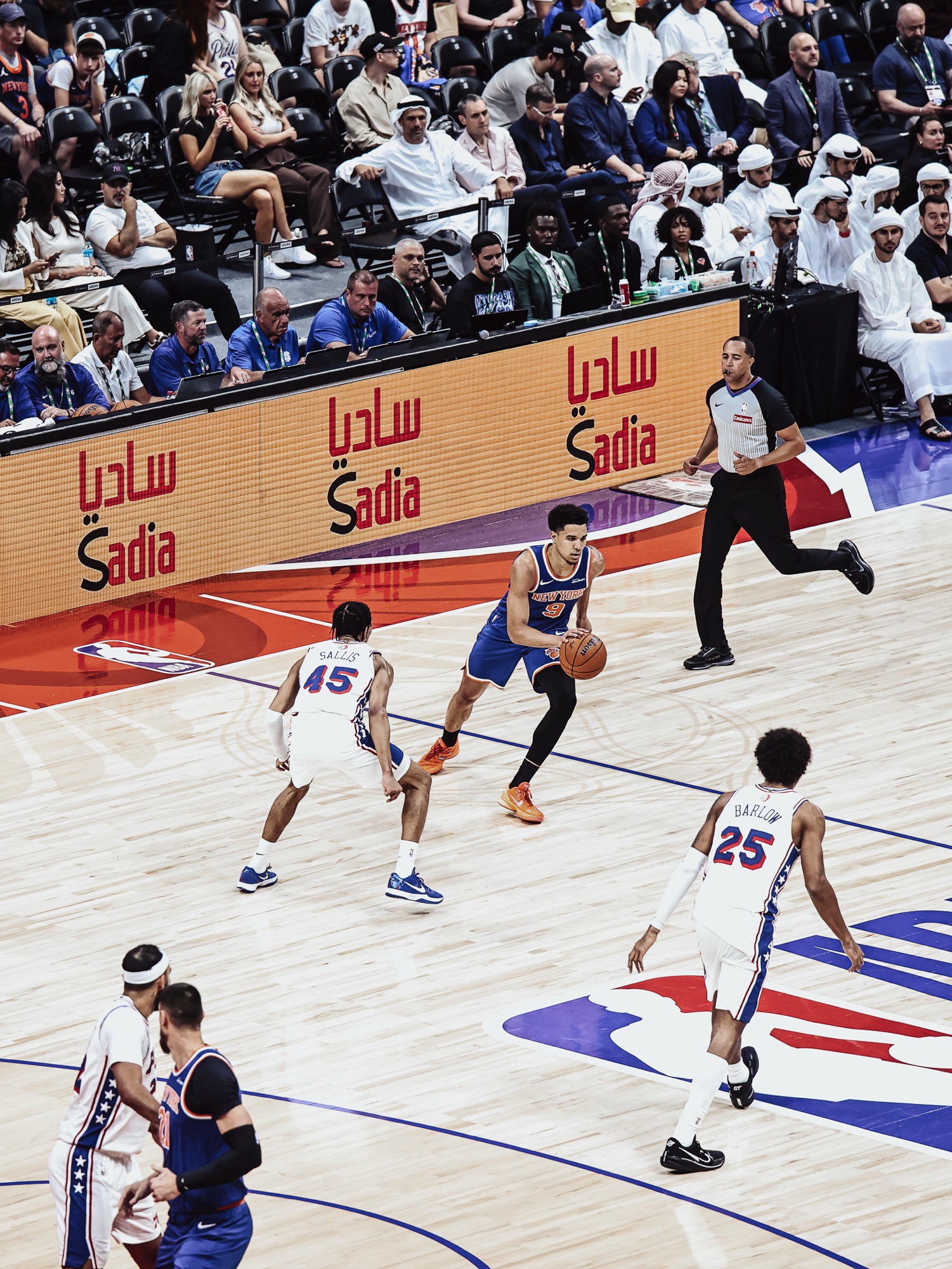 Player in a blue New York jersey dribbling past a defender in front of courtside fans