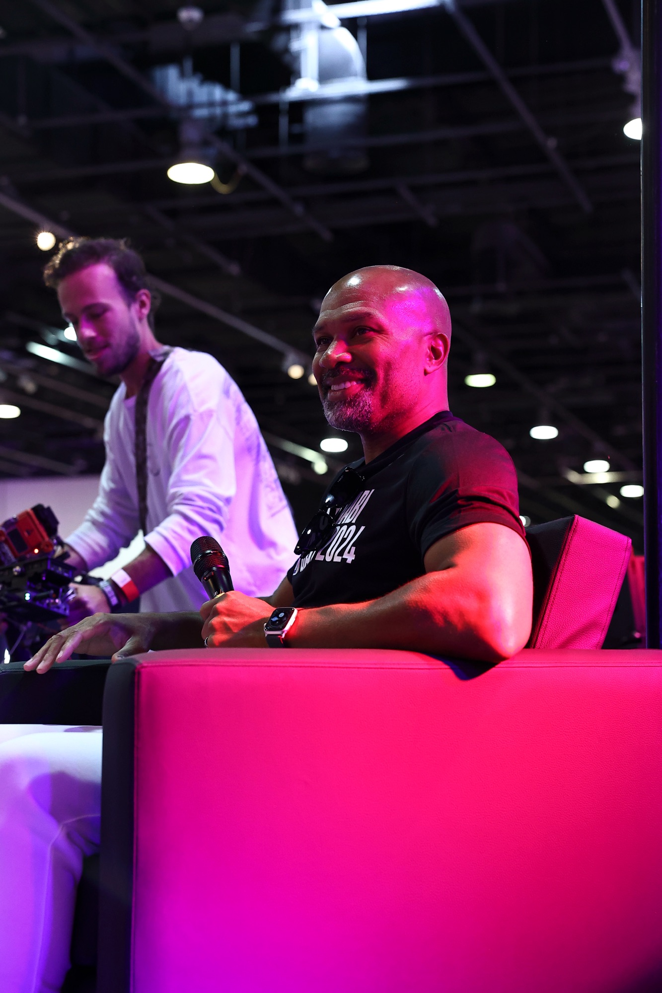 Smiling man holding a microphone on a pink sofa under event lighting
