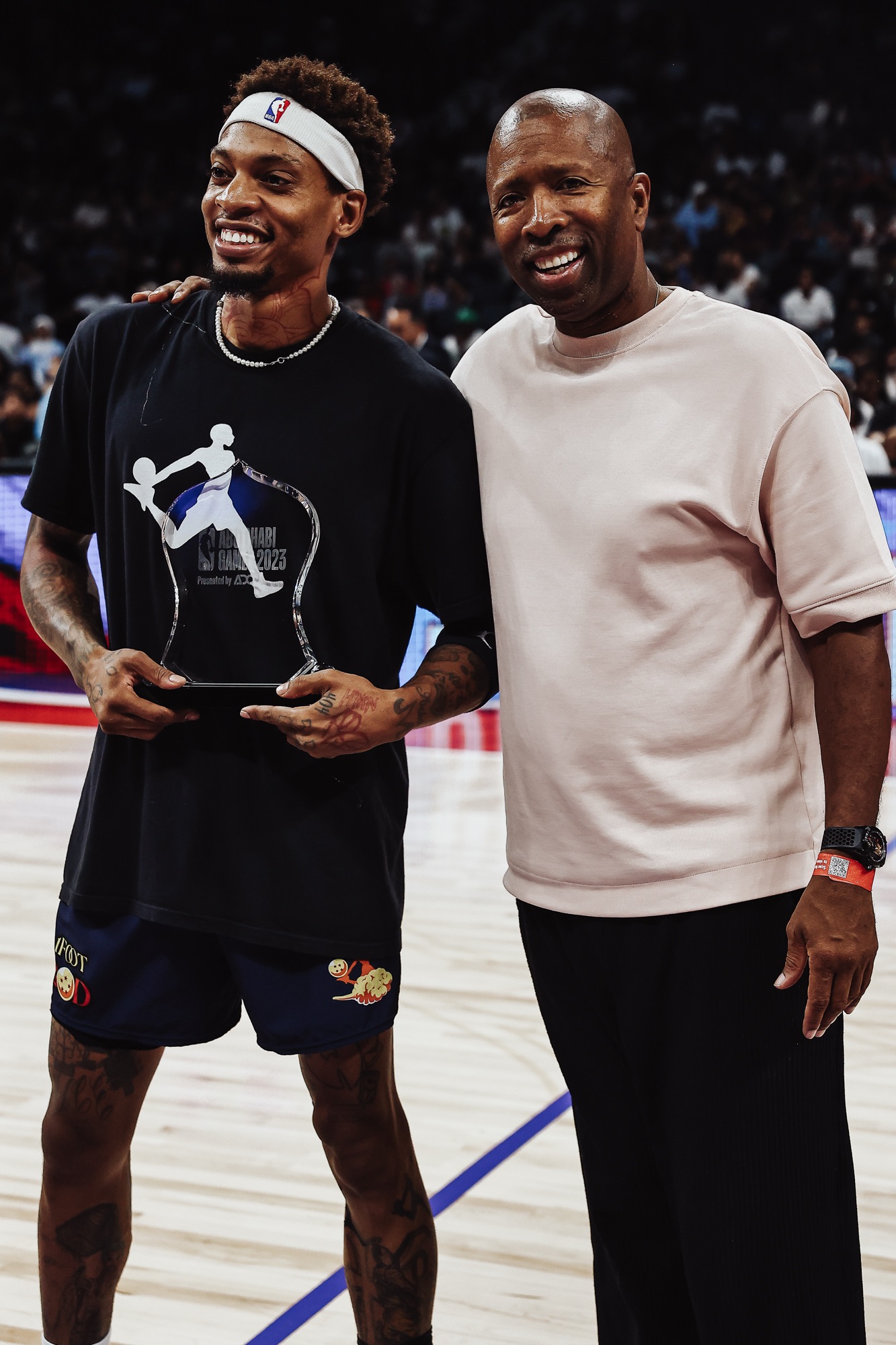Smiling player in a headband holding an award trophy beside a man in a pale pink top on court