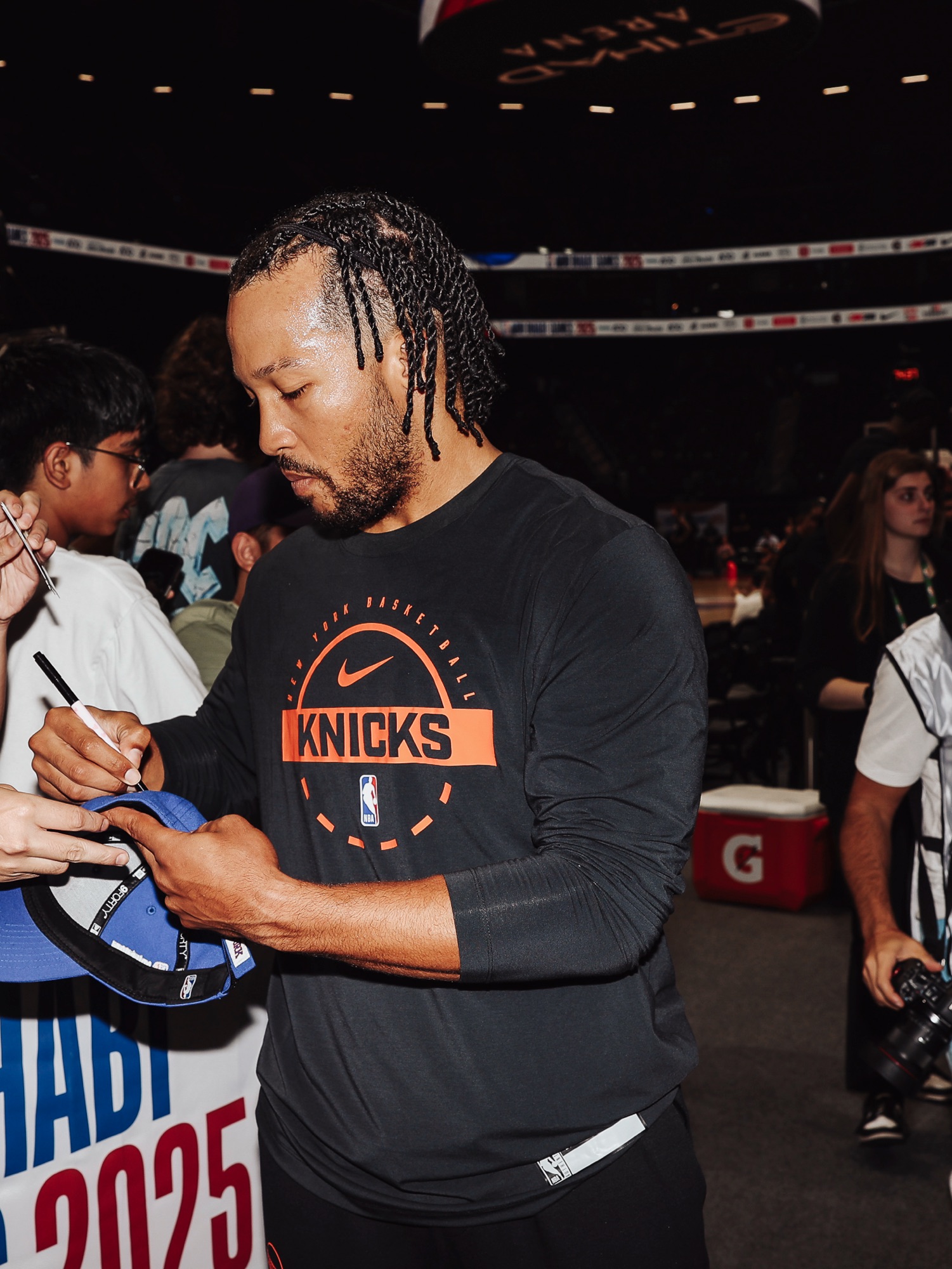 Player in a black Knicks shirt signing a fan's cap courtside