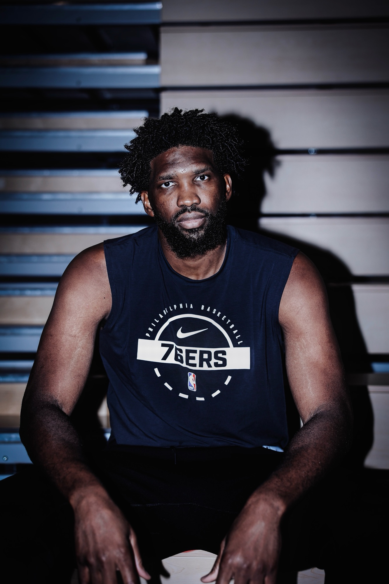 Portrait of a man in a navy 76ers sleeveless top seated on bleachers, moody light