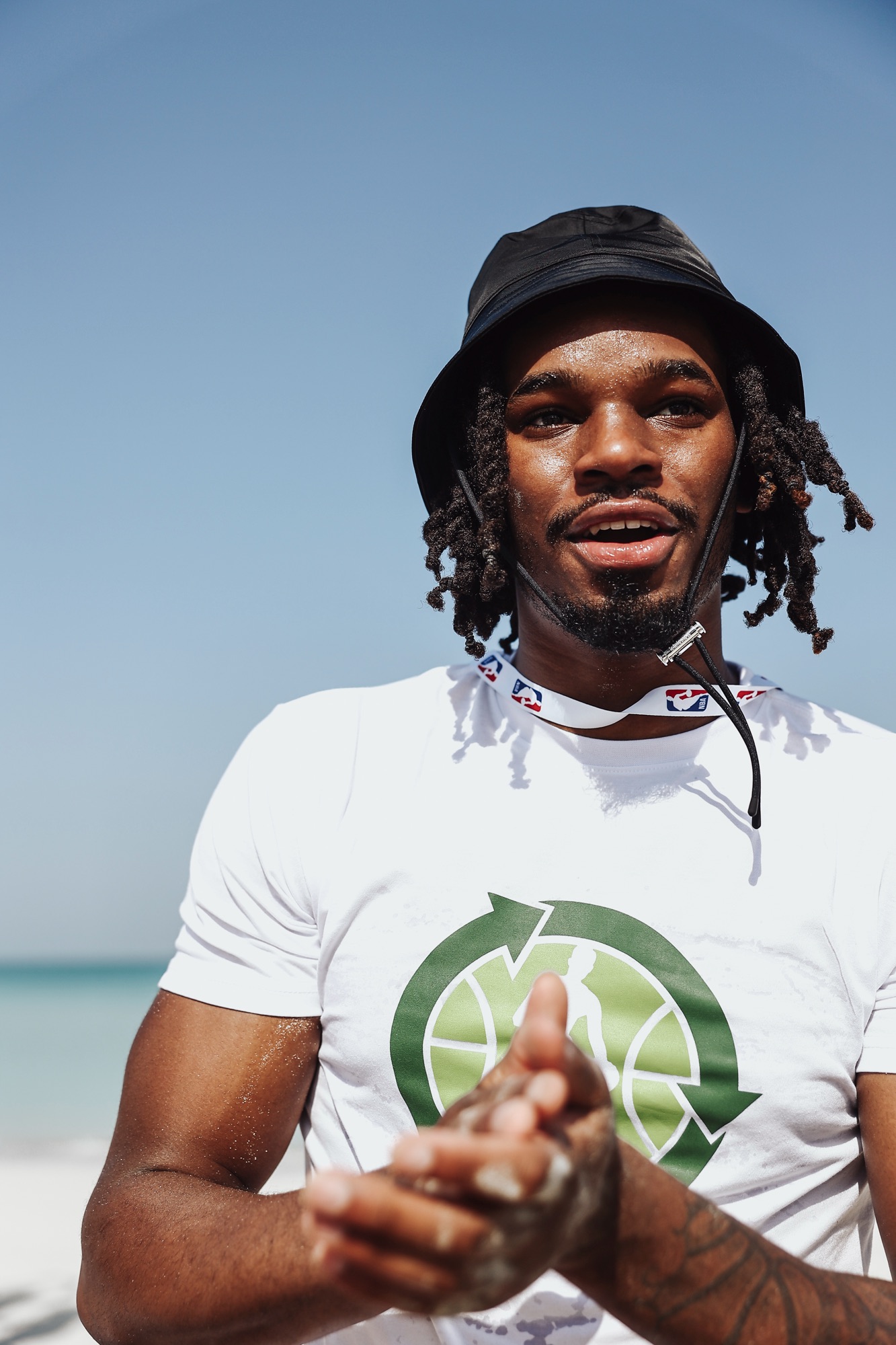 Smiling man in a black bucket hat chatting on the beach in an NBA green T-shirt