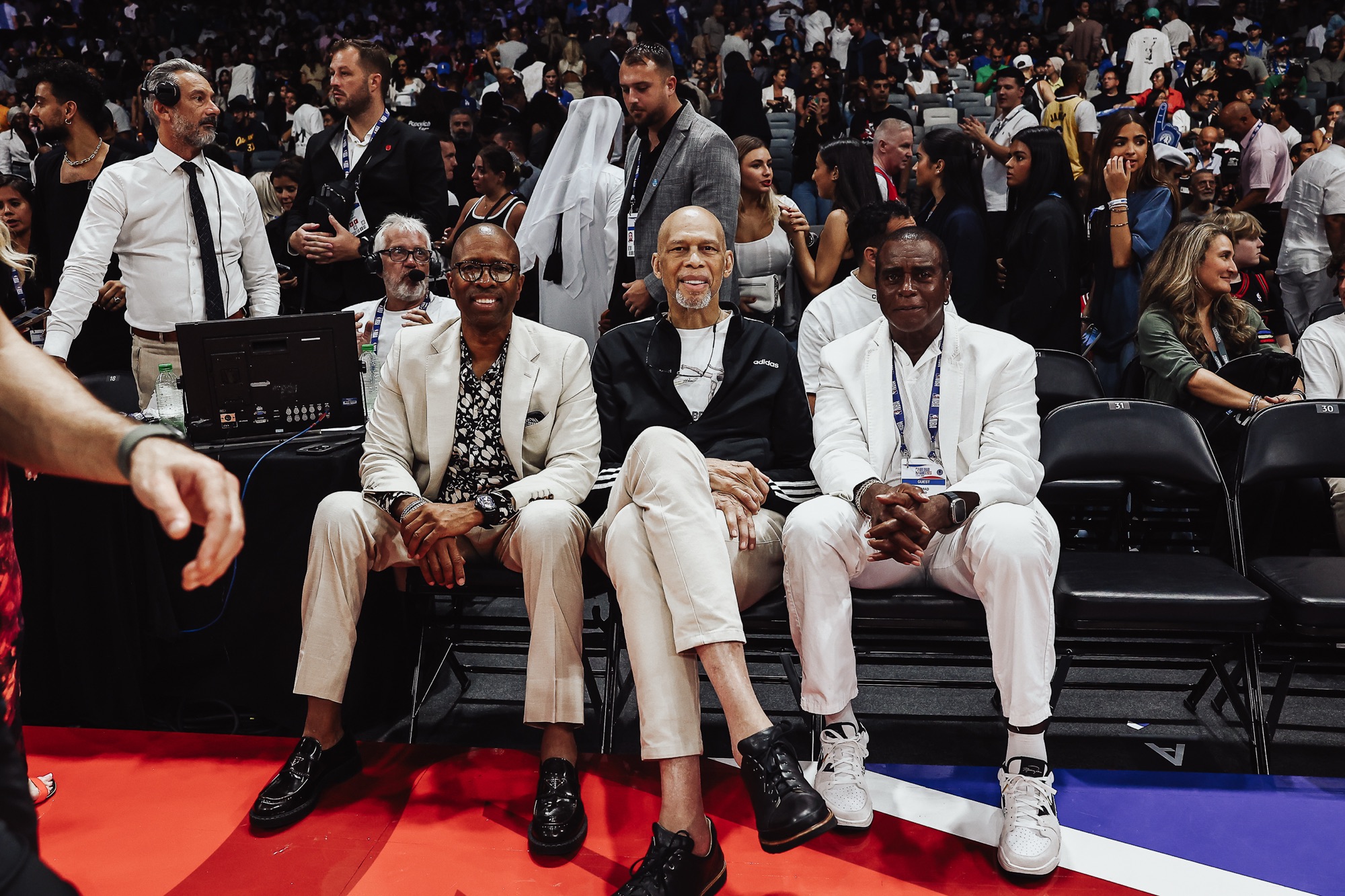 Three men in light suits seated courtside at a packed arena