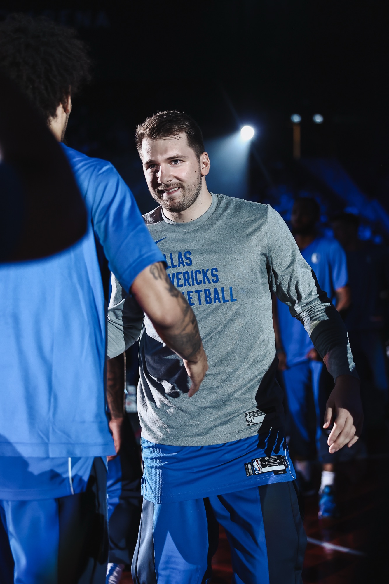 Player in a grey Dallas Mavericks warm-up shirt greeting teammates under spotlights