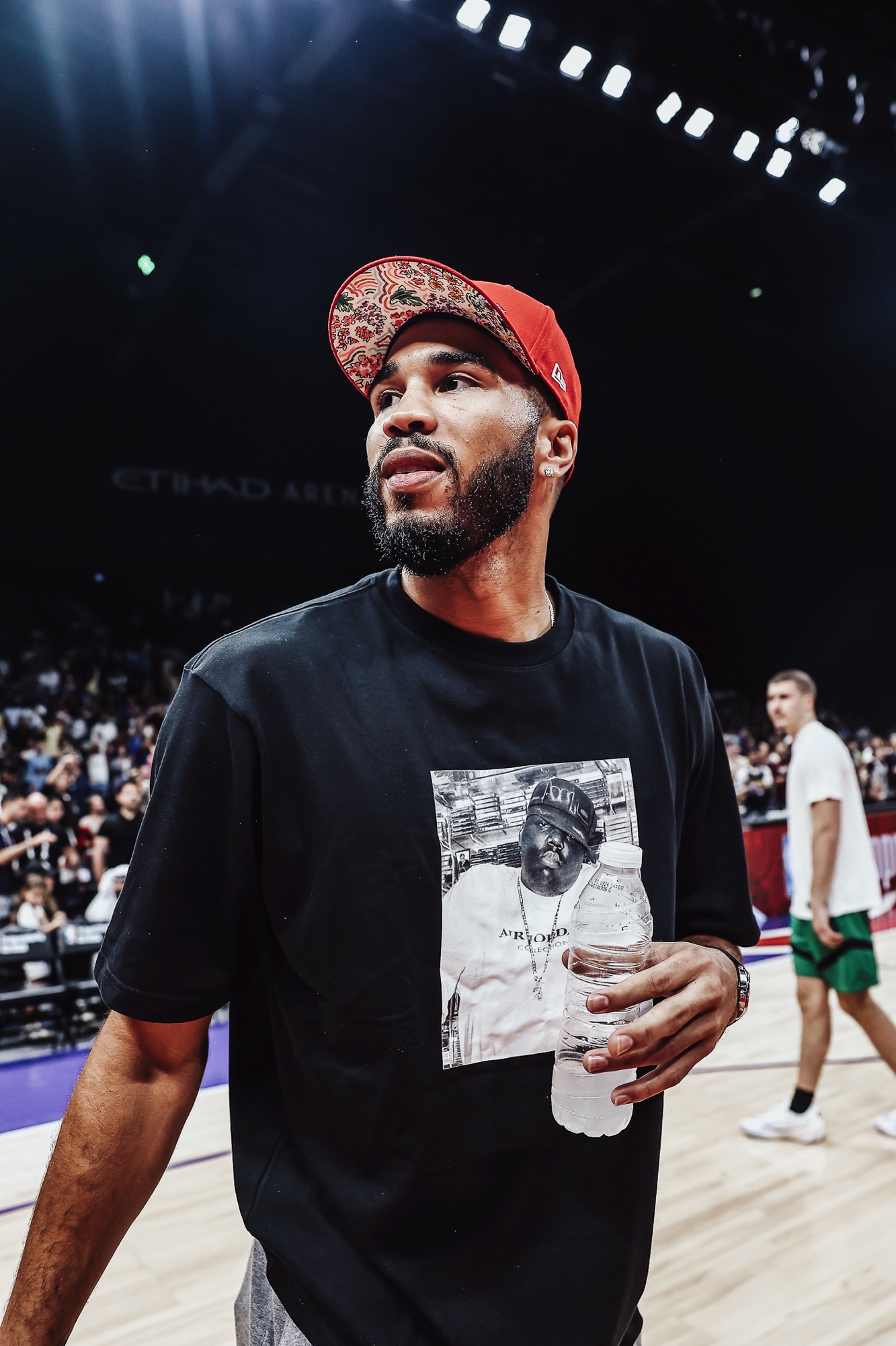 Man in a red patterned cap and graphic T-shirt holding a water bottle courtside