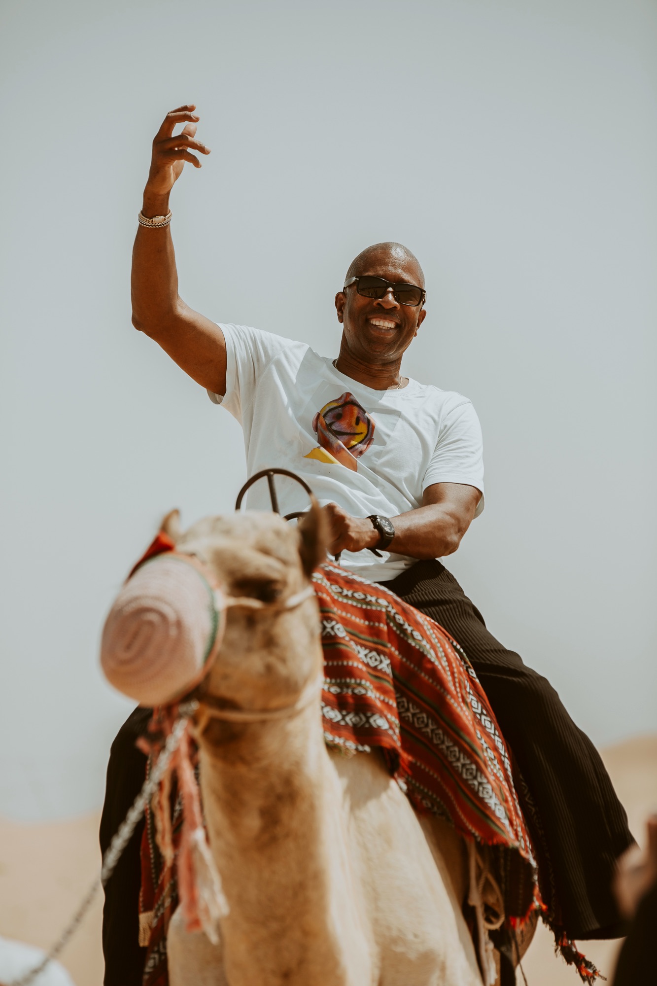Smiling man in sunglasses waving while riding a camel in the desert
