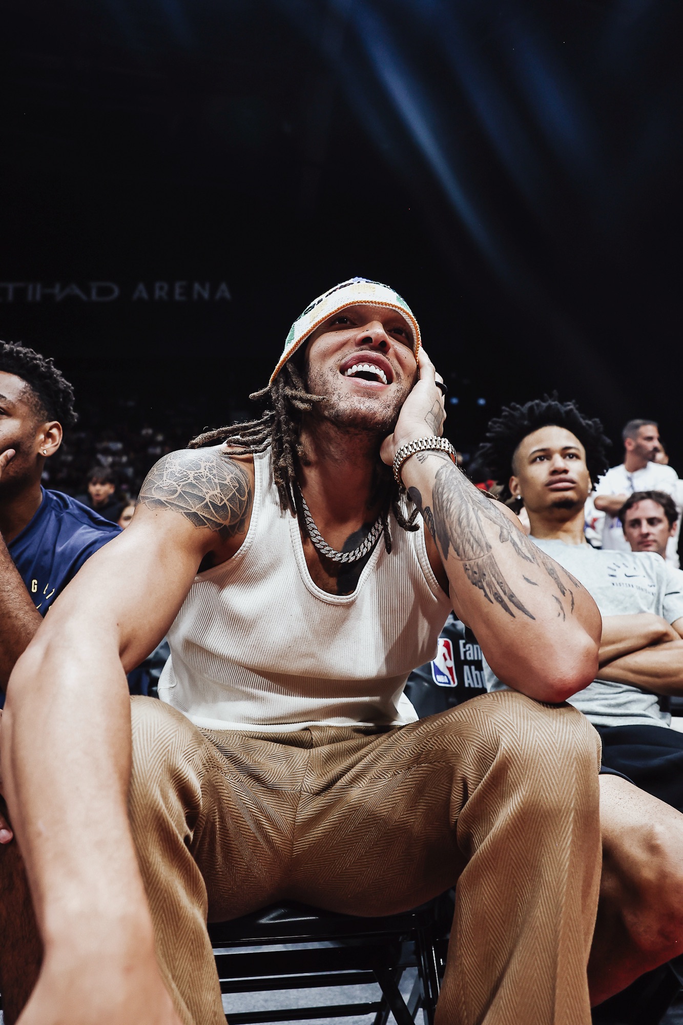 Laughing man in a bucket hat and white tank top sitting courtside at Etihad Arena