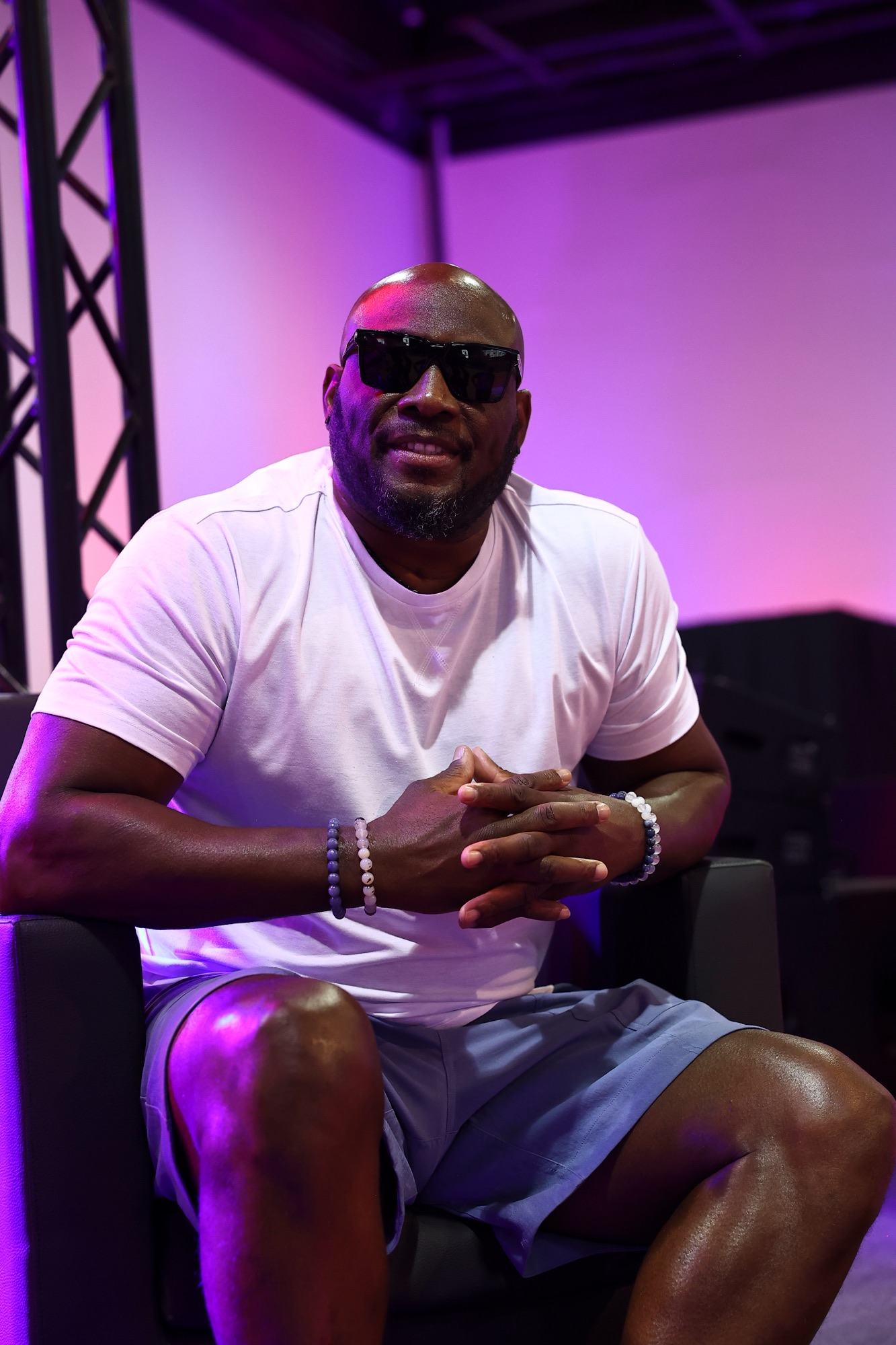 Man in sunglasses and a white T-shirt seated on a pink-lit stage