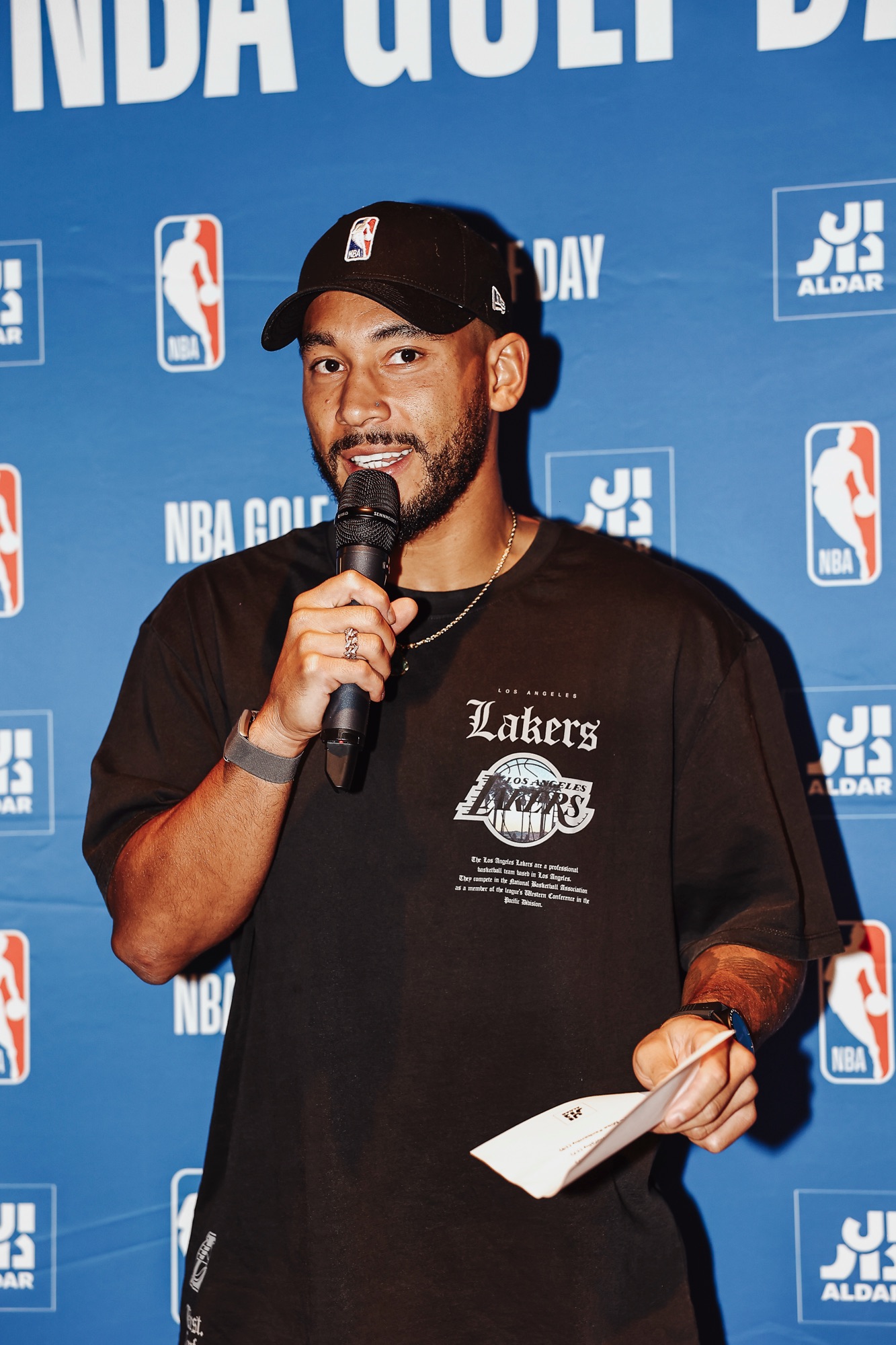Man in a black Lakers T-shirt speaking into a microphone at an NBA Golf Day event