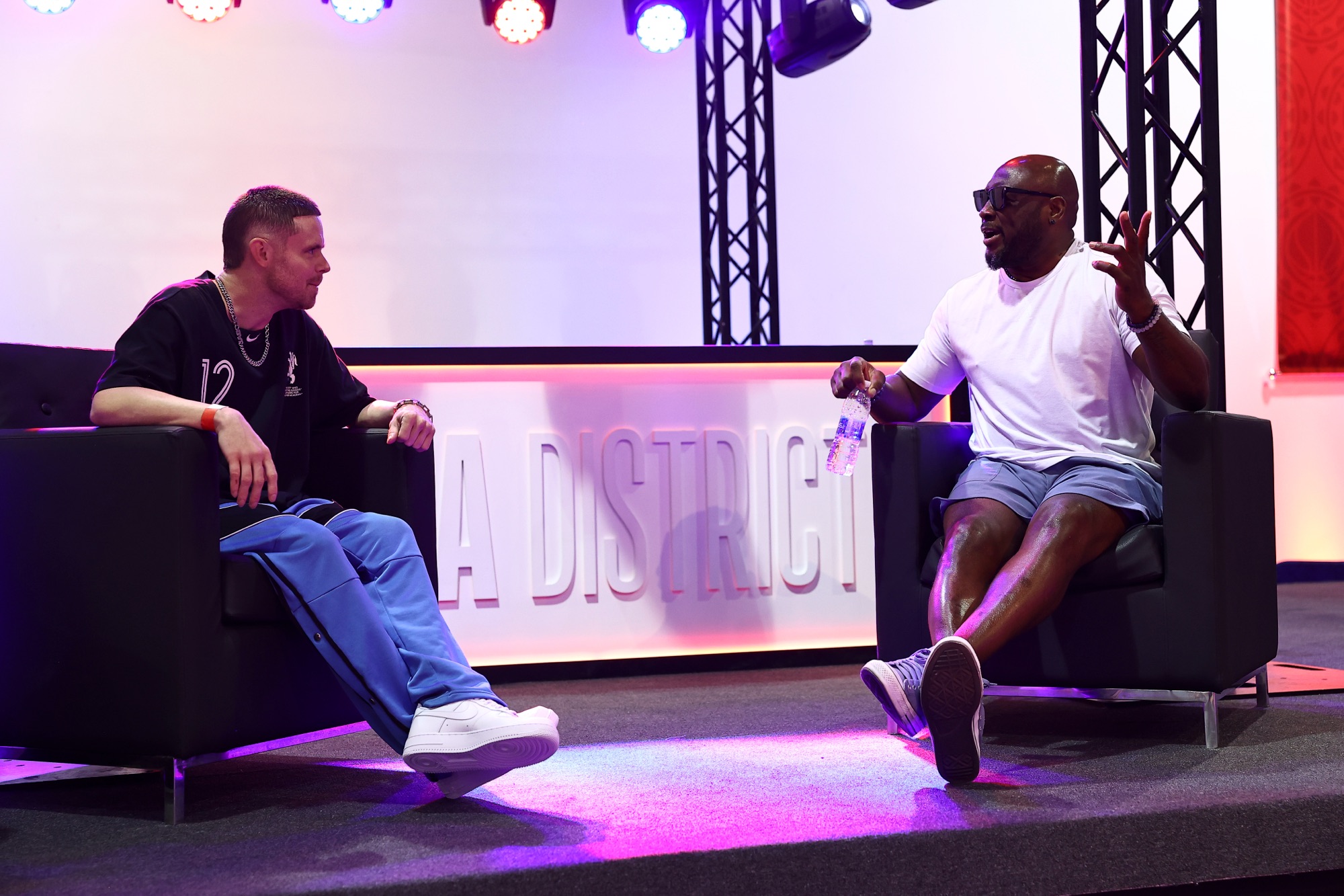 Two men in conversation on armchairs during a stage talk under colourful lights