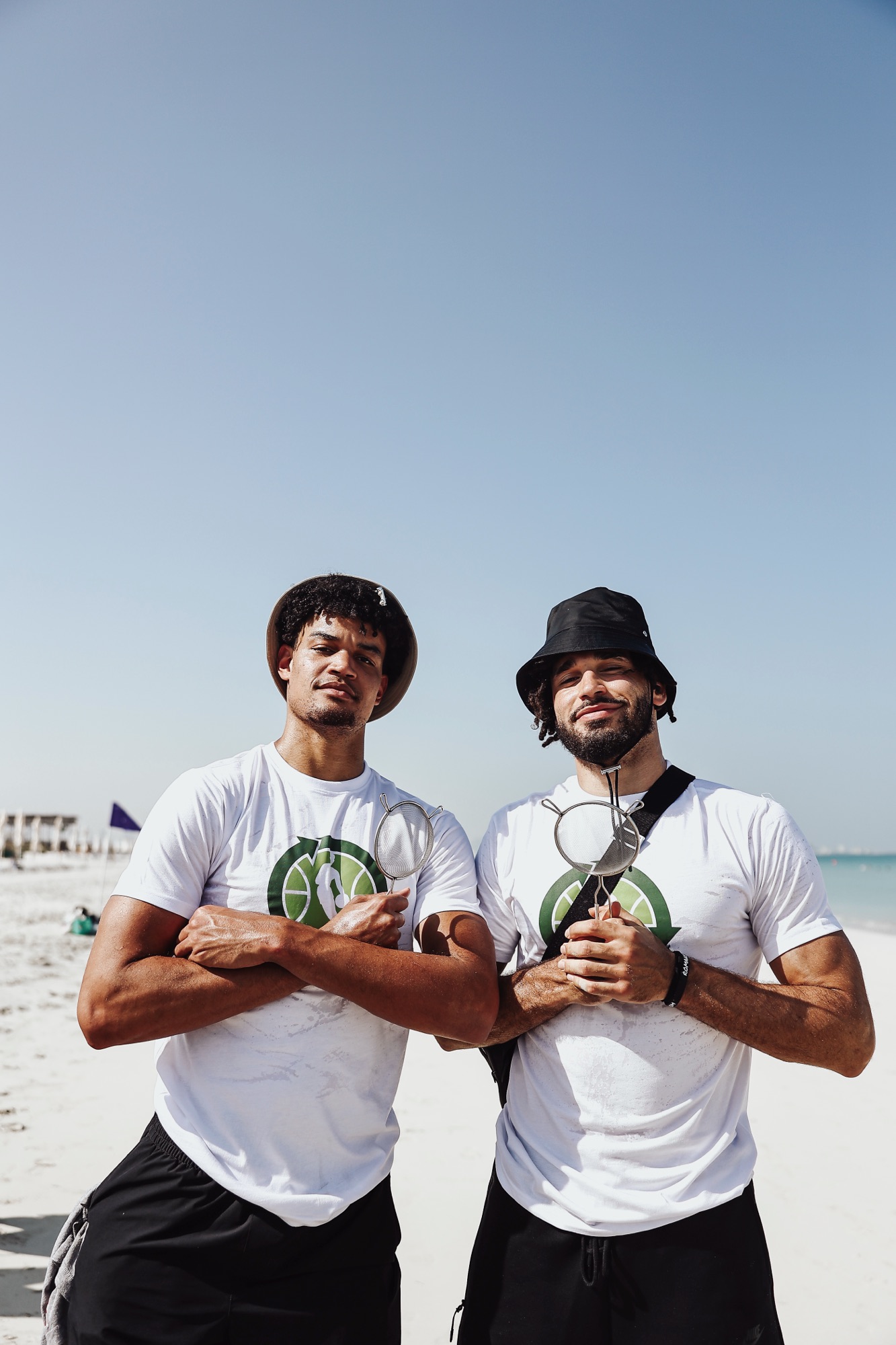 Two men in white T-shirts and hats posing with arms crossed on a sunny beach