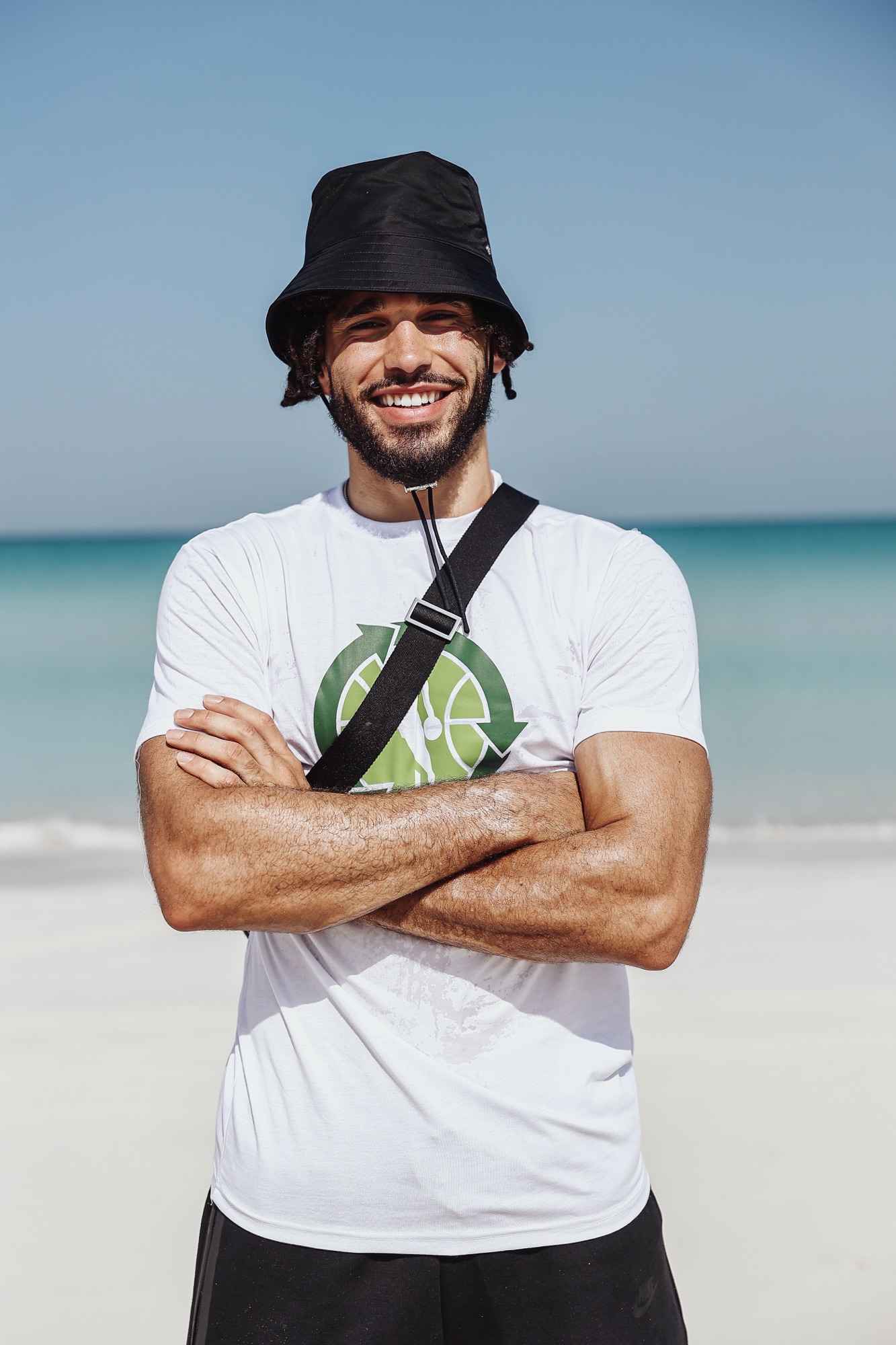 Smiling man in a black bucket hat and white T-shirt on the beach, arms crossed
