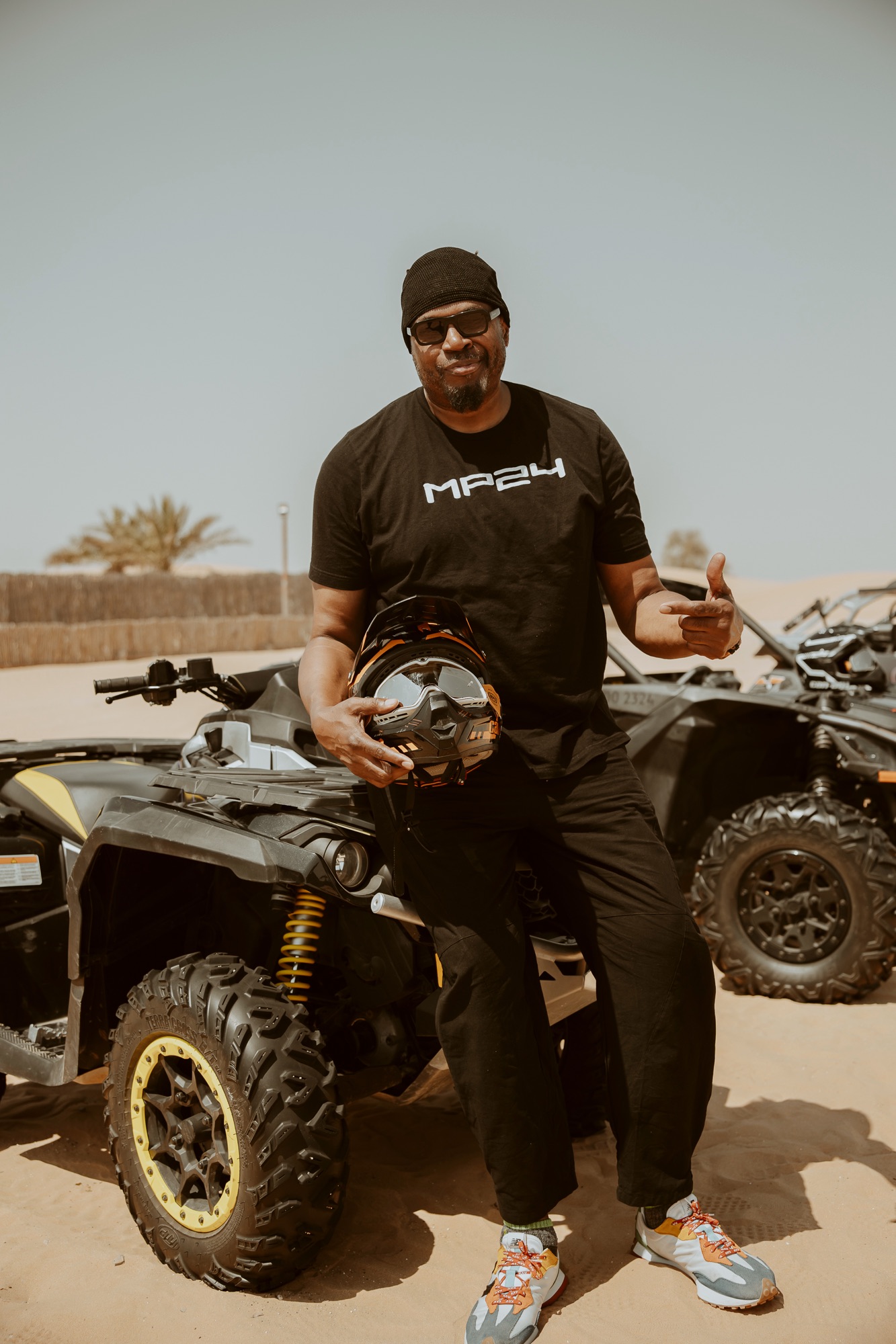 Man in sunglasses holding a helmet beside quad bikes in the desert