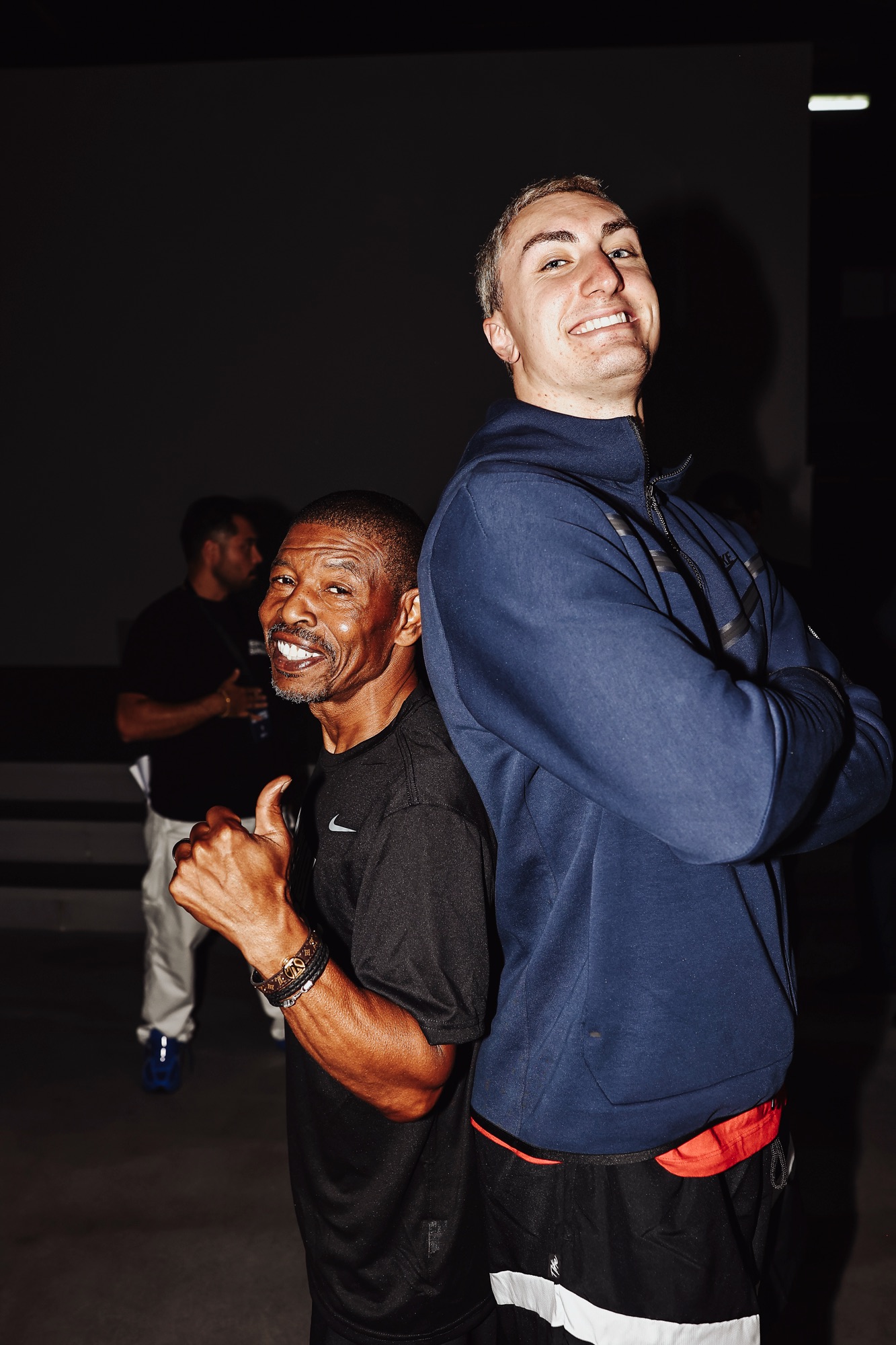 Two smiling men standing back to back, one giving a thumbs-up