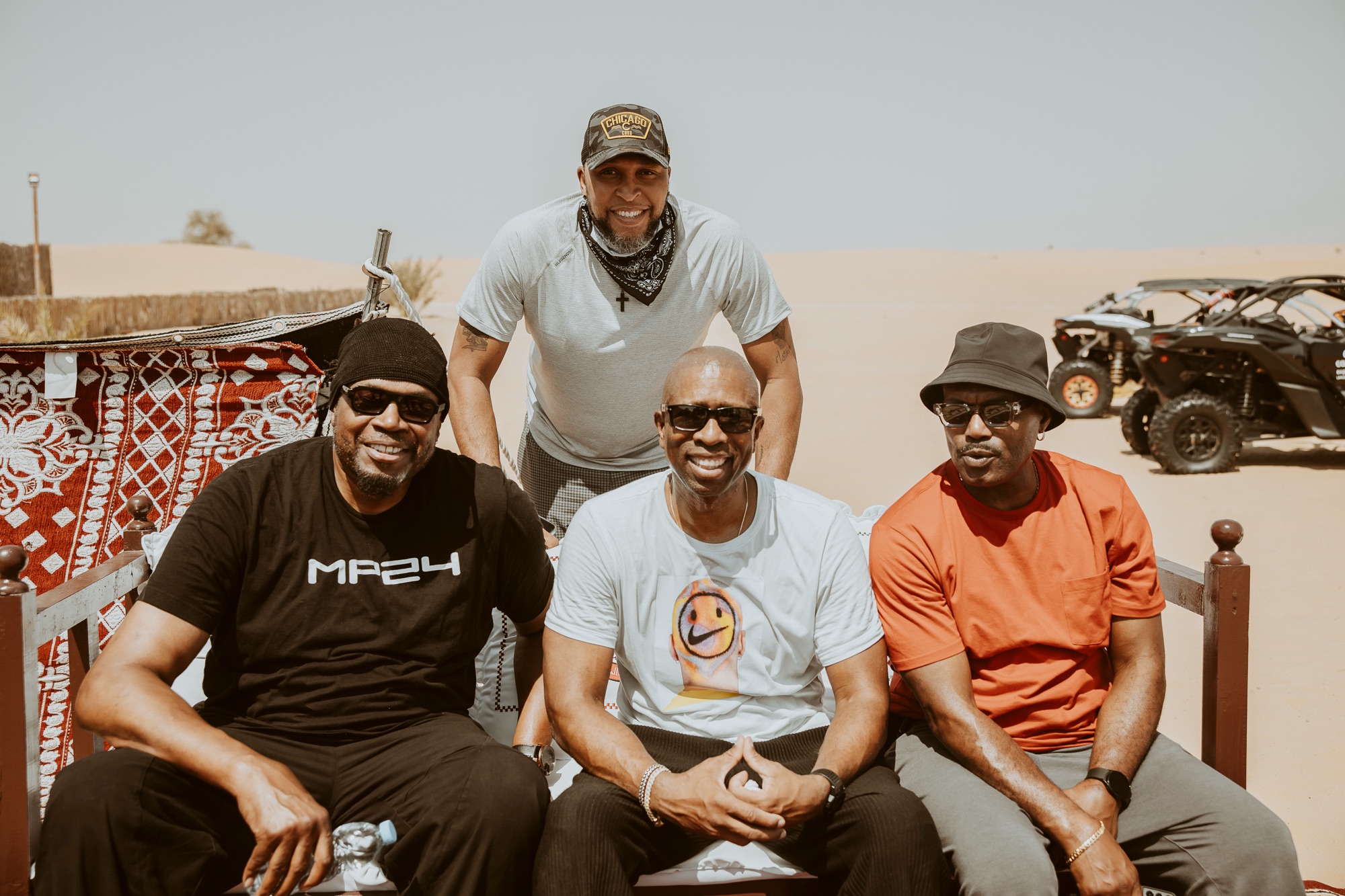 Four smiling men in sunglasses relaxing at a desert camp beside dune buggies