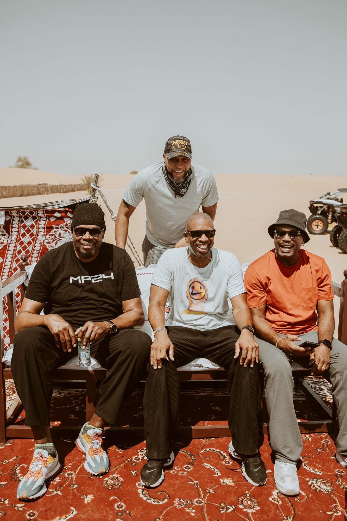 Four men seated on a rug at a desert camp, full-length view