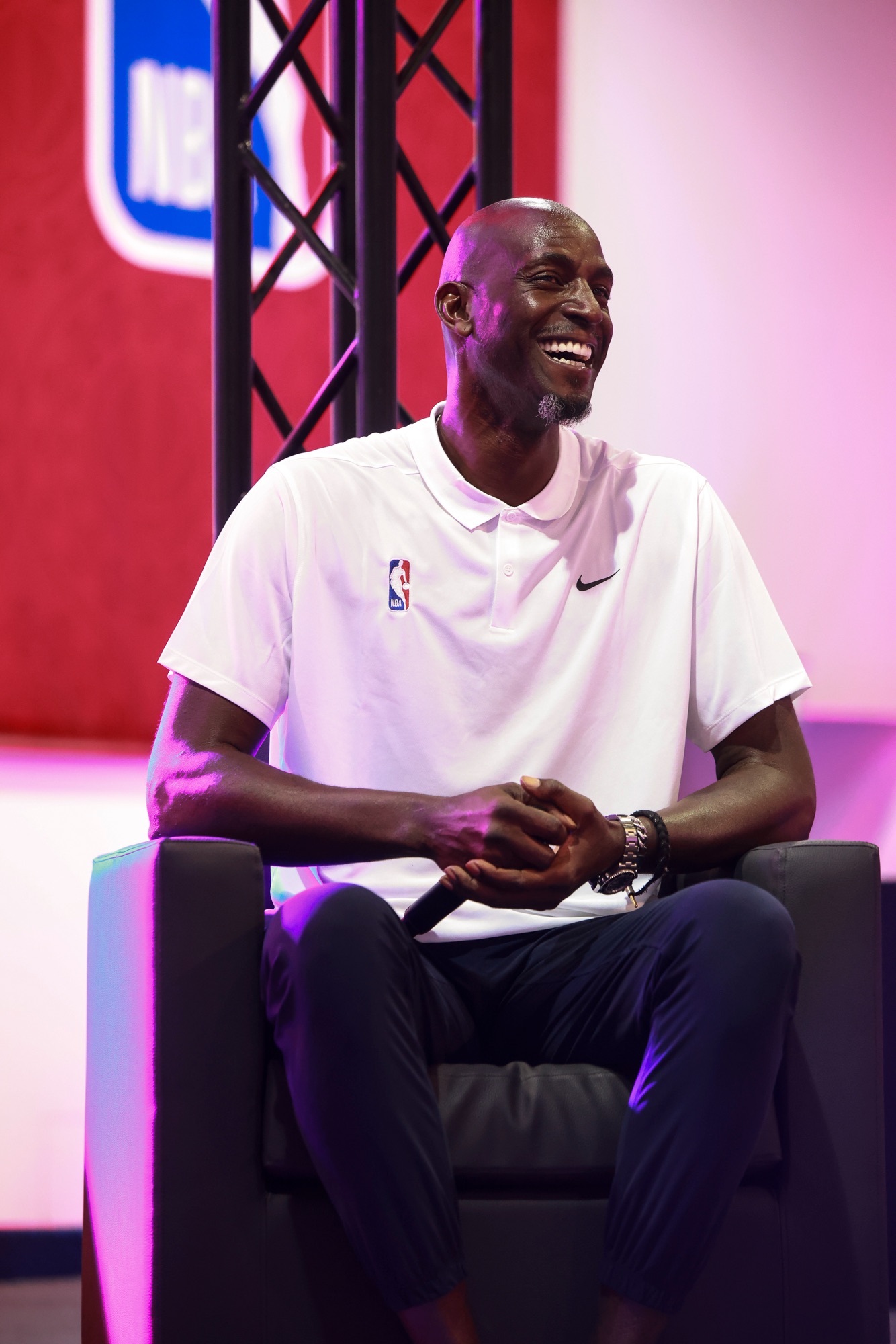 Laughing man in a white NBA polo seated on stage under pink lighting