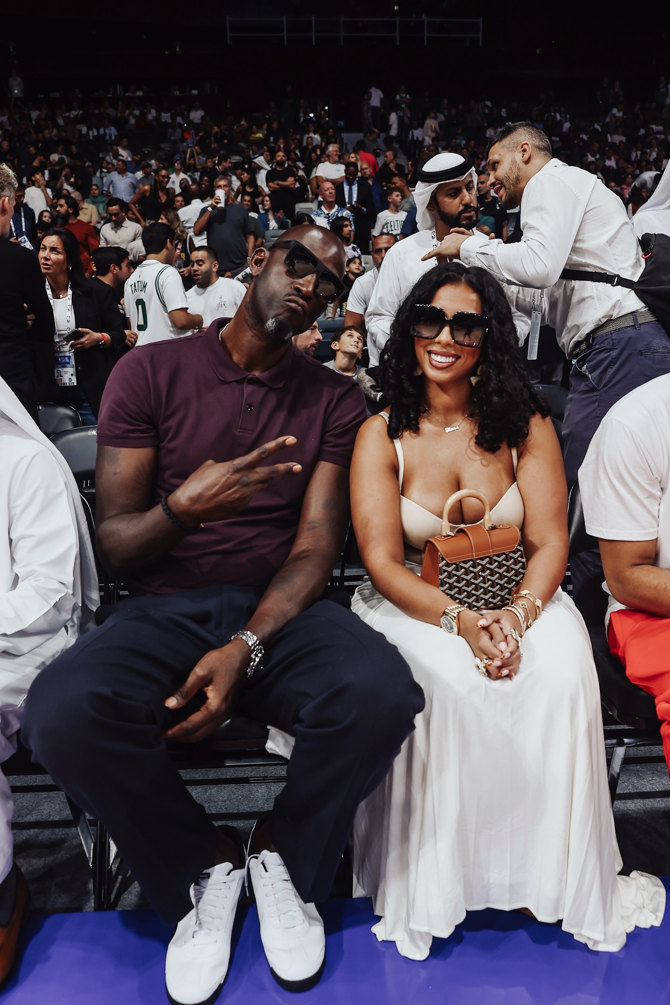 Man in sunglasses making a peace sign beside a smiling woman in white, courtside