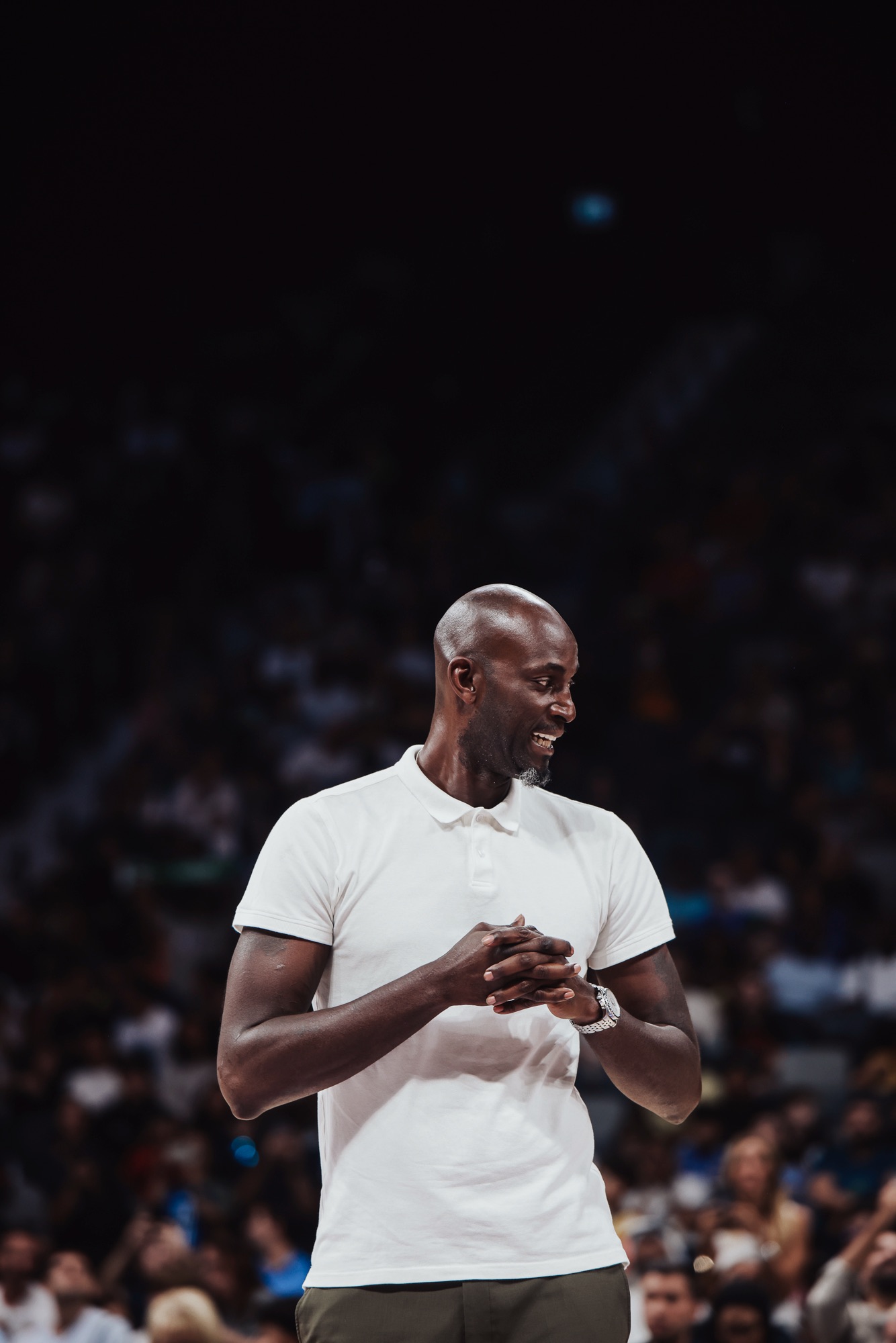 Smiling man in a white polo standing on court against a blurred crowd