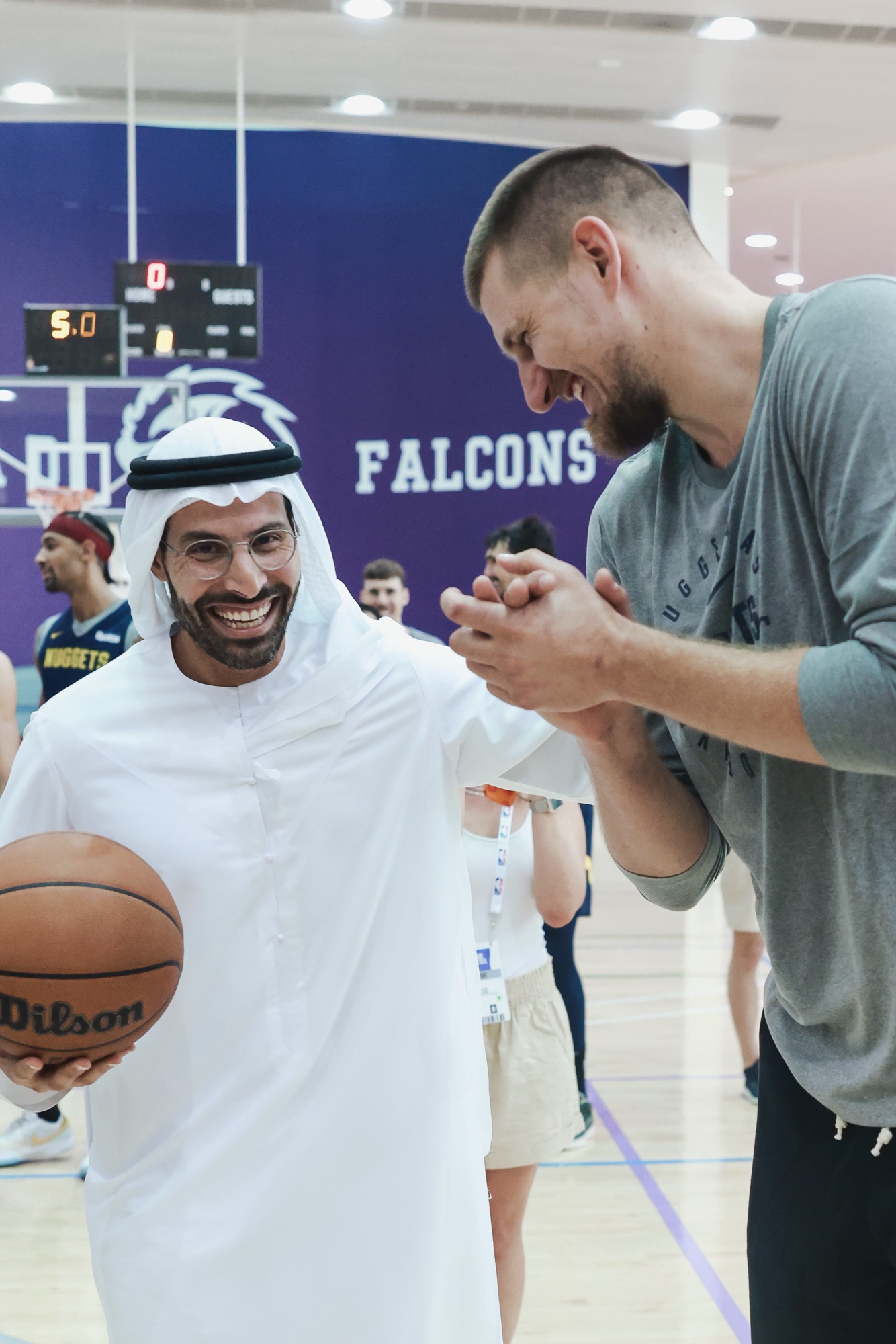 Man in a white kandura holding a basketball and laughing with a player in grey