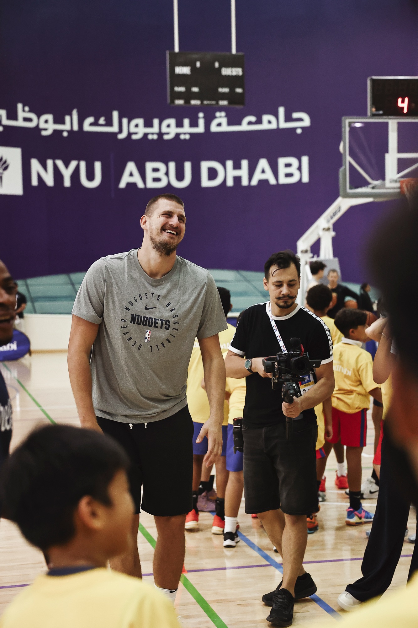 Smiling player in a grey Nuggets T-shirt at a youth clinic as a videographer films