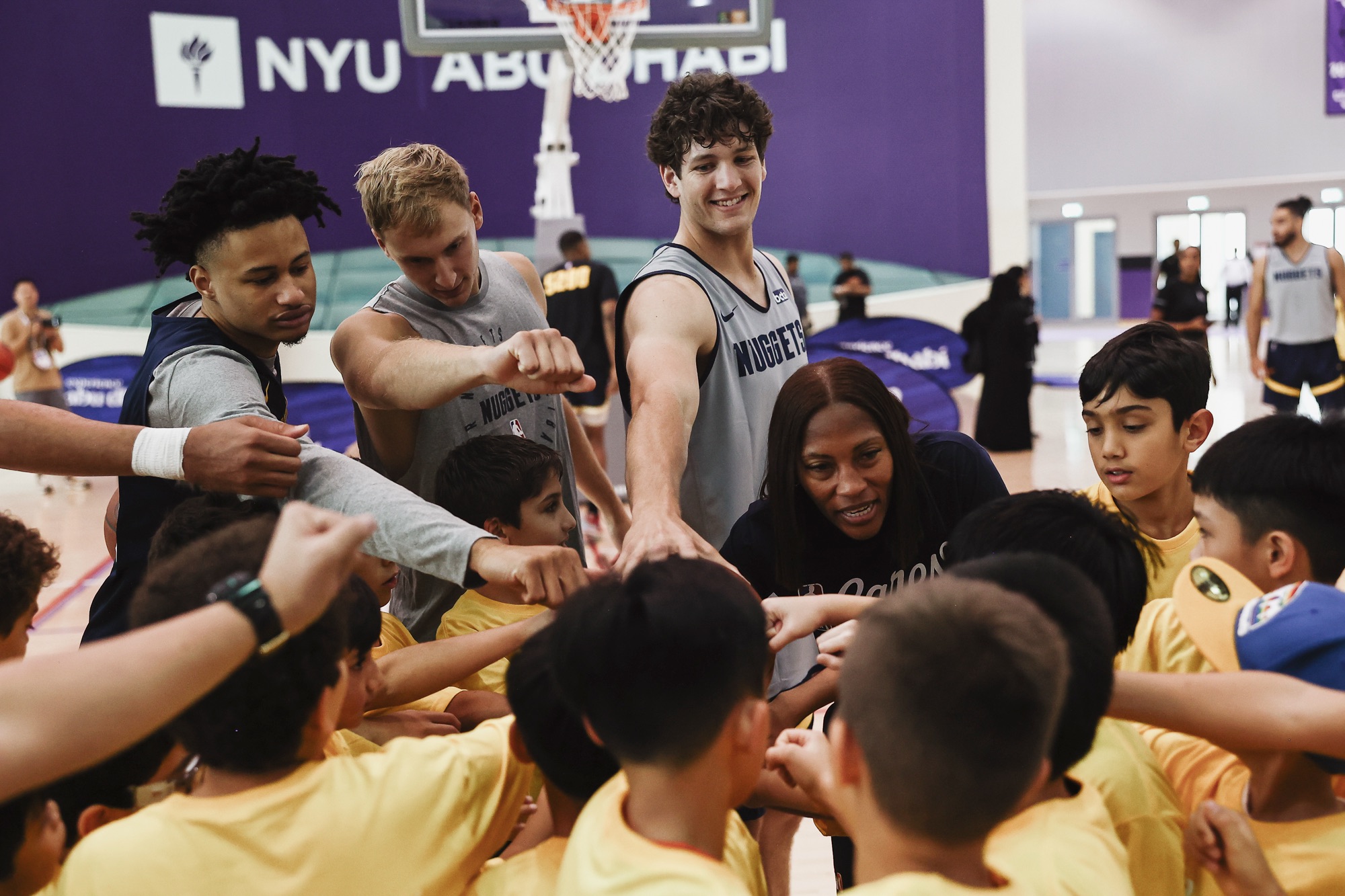 Nuggets players and young campers in yellow joining hands in a team huddle