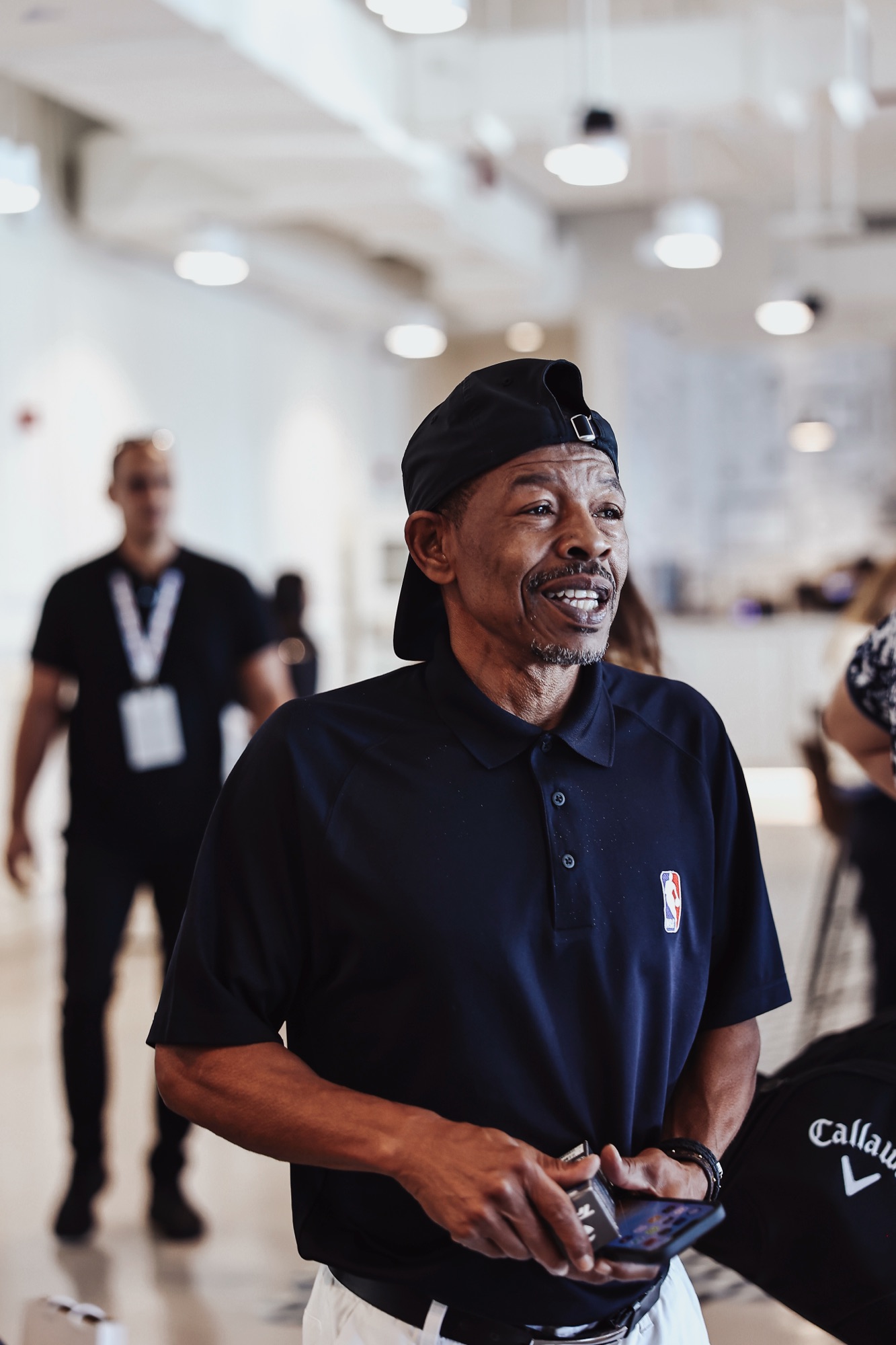 Smiling man in a backwards cap and navy NBA polo at an indoor event